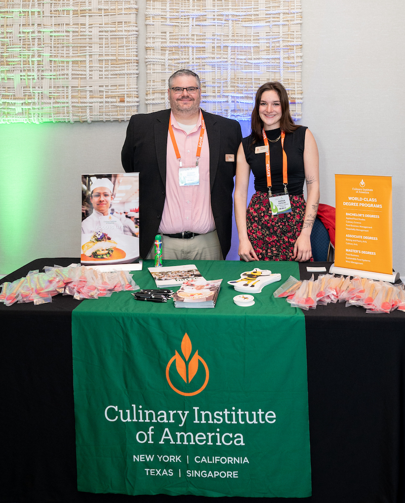 Photo of two representatives from the Culinary Institute of America standing behind their exhibit table at NPSI 2024